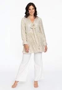 Beige Paillettenoberteil mit V-Ausschnitt, Schleife und langen Ärmeln, kombiniert mit weißen weiten Hosen und sandalen mit Riemchen. Insgesamt glatte Textur.