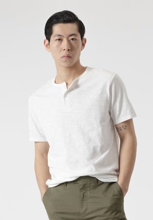 Joven con el pelo negro peinado hacia atrás, lleva una camiseta henley blanca de manga corta y pantalones verde oliva, manos en los bolsillos, expresión neutral.