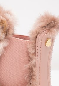 Acercamiento de una bota forrada de piel de color rosa con un panel textil de punto y un pequeño emblema decorativo dorado en el lado exterior liso.