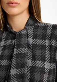 Grau-schwarz kariertes Material mit weicher Textur, ausgestattet mit einem hohen Kragen und einem Knoten-Detail am Ausschnitt. Nahaufnahme des Designs.