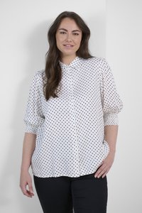 Blouse blanche à pois noirs, avec un devant boutonné, un col et des manches courtes bouffantes. Tissu lisse et coupe décontractée. Pantalon noir porté.