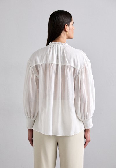 Blouse blanche semi-transparente avec des manches bouffantes et un col froncé. Associée à un pantalon de couleur claire. Texture douce avec une légère transparence.