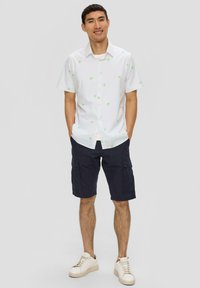 Weiße Kurzarmbluse mit grünem Palmenmuster, Knopfleiste vorne, kombiniert mit navy Cargo-Shorts und weißen Turnschuhen.