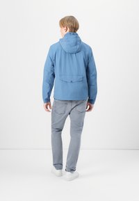 Lyseblå jakke i lett materiale med hette, elastiske mansjetter og baklomme. Kombinert med grå jeans og hvite joggesko.