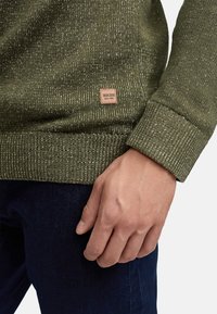Oliwkowy sweter dzianinowy z teksturowanym wykończeniem, z skórzanym metkiem oraz ściągaczami przy mankietach i u dołu. Ręka oparta na tkaninie.