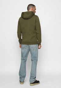 Sudadera verde oliva con capucha ajustable, puños y dobladillo de canalé, combinada con jeans de mezclilla azul claro y zapatillas negras.