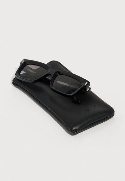 Μαύρα ορθογώνια γυαλιά ηλίου που ακουμπούν σε μια μαλακή μαύρη δερμάτινη θήκη πάνω σε απλό λευκό φόντο.
