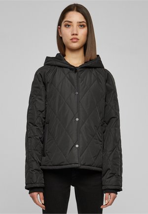 Femme portant une veste matelassée noire à capuche avec des boutons-pression, debout devant un fond uni gris clair.