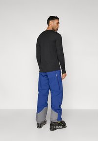 Crna košulja dugih rukava u kombinaciji s plavim skijaškim hlačama koje imaju sive detalje, patentne zatvarače sa strane i crne skijaške čizme.