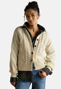 Krémový kardigán s pleteným vzorom a rebrovaným lemom, prehodený cez modrú károvanú košeľu. Obsahuje textúrované vzory a objemné rukávy.