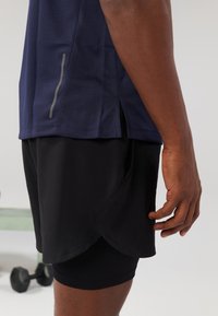 Maglietta sportiva navy in tessuto testurizzato, con aperture laterali e accenti riflettenti, abbinata a pantaloni corti neri. Materiale liscio, design aderente.