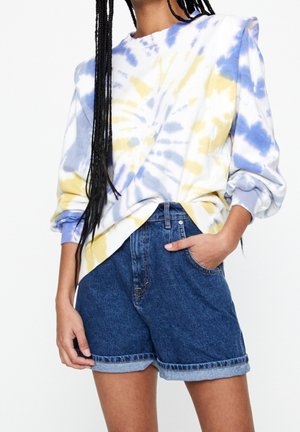 Person wearing blue and yellow tie-dye sweatshirt with rolled sleeves and high-waisted rolled-hem denim shorts, hand in pocket.