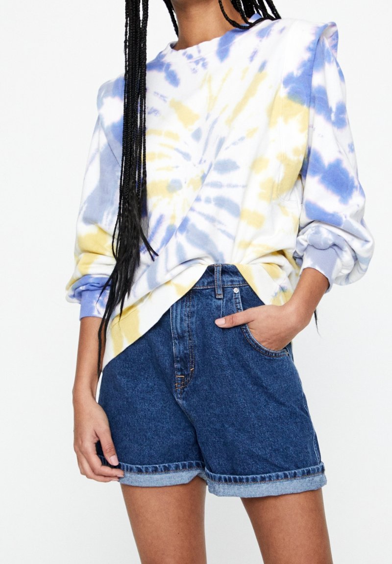 Personne portant un sweat-shirt tie-dye bleu et jaune avec manches retroussées et short en jean taille haute à ourlet roulotté, main dans la poche.