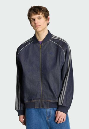 Ung mand iført en mørk denim lynlåstrøje med stribede ærmer og blå jeans, stående mod en ensfarvet lys baggrund.