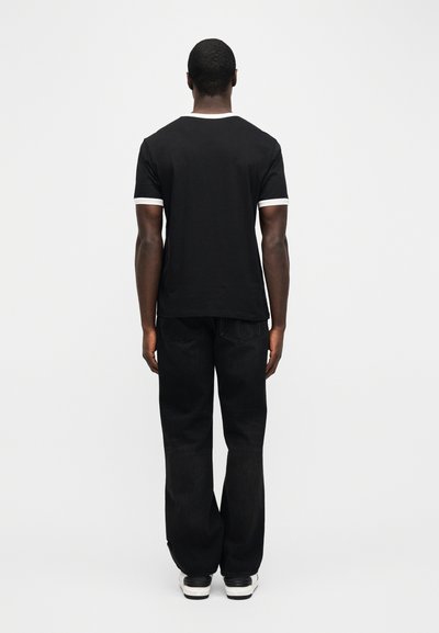 Άνδρας που στέκεται με την πλάτη γυρισμένη, φορώντας μαύρο μπλουζάκι με λευκή περιφέρεια, μαύρα τζιν και λευκά αθλητικά παπούτσια σε απλό λευκό φόντο.