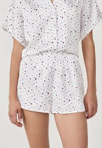 Conjunto de pijama blanco de dos piezas con mangas cortas y pantalones cortos, decorado con pequeños patrones de corazones en rosa, azul marino y burdeos.