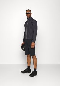 Schwarze leichte Jacke mit Stehkragen, grauem Reißverschluss und reflektierenden Akzenten. Schwarze Shorts und Sportschuhe, ein schwarer Helm in der Hand.