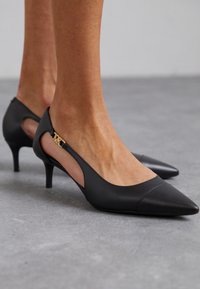 Talons en cuir noir avec des bouts pointus et des côtés découpés. Accent doré MK sur le talon. Talon slim de style stiletto de 5 cm. Texture lisse.
