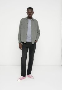 Γκρι πουκάμισο με κουμπιά πάνω από ανοιχτό γκρι μπλουζάκι, μαύρο τζιν και ροζ αθλητικά παπούτσια. Ματ υφή, καθημερινό σχέδιο και τσέπες στο πουκάμισο.