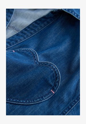 Džinsa auduma krekls zilā tonī ar sirdsdizaina kabatu, kas izšūta rozā krāsā. Gluda tekstūra, ikdienišķs dizains.