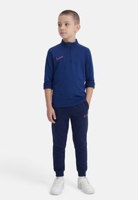 Νεαρός αγόρι που φοράει μπλε σκούρο φούτερ με φερμουάρ και παντελόνι με ροζ λογότυπα Nike, λευκά αθλητικά παπούτσια Nike, κοιτάζει προς το πλάι με τα χέρια στις τσέπες.