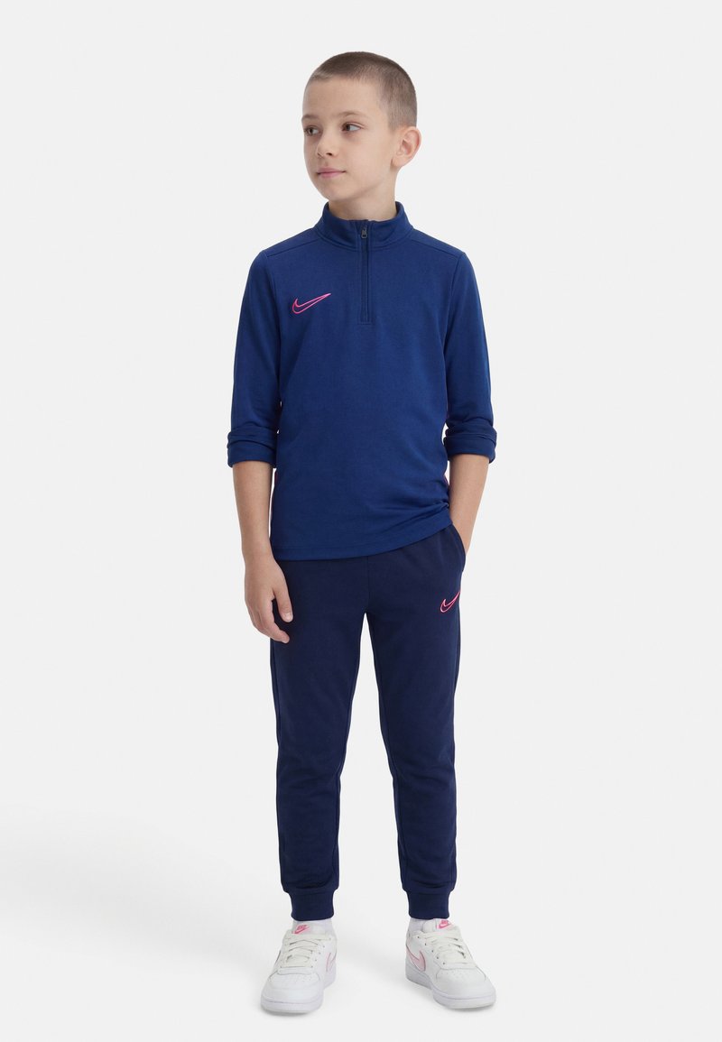 Νεαρός αγόρι που φοράει μπλε σκούρο φούτερ με φερμουάρ και παντελόνι με ροζ λογότυπα Nike, λευκά αθλητικά παπούτσια Nike, κοιτάζει προς το πλάι με τα χέρια στις τσέπες.