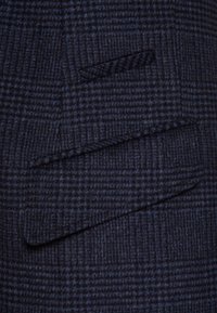 Granatowa marynarka w kratkę z teksturowanej mieszanki wełny. Posiada wywijany kołnierz oraz dwie skośne kieszenie. Subtelne detale w wzór jodełki.