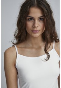 Weißes Tanktop mit dünnen Trägern, glatter Textur und tailliertem Schnitt. Das Model hat lange, wellige Haare und einen neutralen Hintergrund.