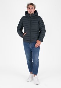Jonge man staand in een zwart gewatteerd jack met capuchon, blauwe jeans en witte sneakers tegen een eenvoudige witte achtergrond.
