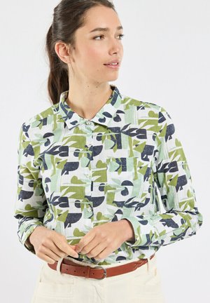 Femme aux cheveux bruns attachés portant une chemise à manches longues à boutons avec des motifs abstraits de feuilles vertes, bleu marine et blanches, en train d'attacher sa ceinture.