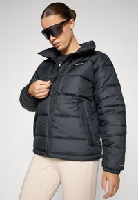 Zwarte pufferjack met een hoge kraag, ritszakken en een gestikt ontwerp. Het model draagt een oversized zwart zonnebril en beige broek.