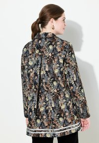 Pitkä kukkakuosiset takki mustasta kankaasta, jossa on vihreän, beige ja oranssin sävyjä. Sisältää teksturoidun helman ja muotoillun kauluksen.