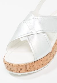 Höyhenmetallinen sandaali, jossa on ristiin menevä yläosa, korkkipohja ja valkoinen pohja. Sileä pinta, jossa on näkyviä ompelusaumoja.