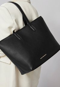 Schwarze Leder-Tote mit einer strukturierten Oberfläche, doppelten Griffen und einem goldenen Markenlogo auf der unteren Vorderseite. Strukturierte Form.