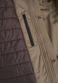 Gewatteerde jas met een beige buitenkant en bruine doorgestikte panelen. Voorzien van een ritszak, drukknopen en een gladde textuur met contrasterende stiksels.