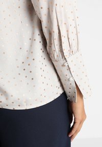 Blusa beige chiaro con pois rosa oro lucido, polsini aderenti e dettagli di maniche plissettate. Tessuto liscio con dettagli in bottoni.