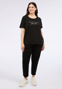 Top negro de manga corta con el texto "Ce la farò anche" y acentos en verde y rojo, combinado con pantalones negros y zapatillas blancas. Diseño simple y casual.
