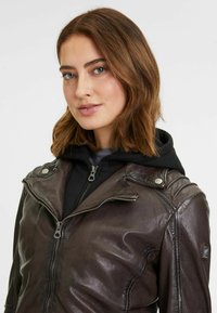 Giubbotto biker in pelle marrone con chiusura a zip, bottoni a pressione e dettagli sulle spalle. Indossato sopra un felpa nera con cappuccio.