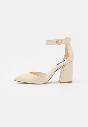 Tacon de charol beige con correa al tobillo, punta afilada, tacón bloque y hebilla ajustable. Textura suave con acabado brillante.