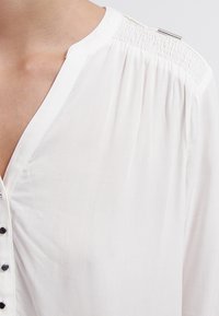 Blouse blanche en tissu léger, avec un décolleté en V profond, une fermeture à boutons et des détails froncés aux épaules ornés d'accents métalliques.