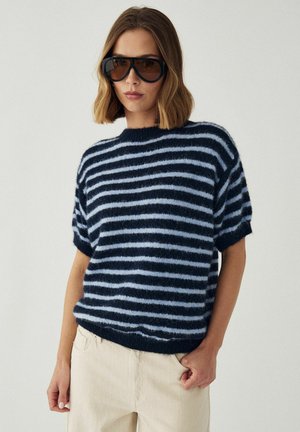 Kvinde iført store mørke solbriller og en sort- og lyseblå stribet fluffy kortærmet sweater med beige bukser.