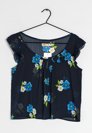 Marineblauwe blouse met korte geplooide mouwen, versierd met blauwe en witte bloemmotieven, hangend aan een zwarte kledinghanger.