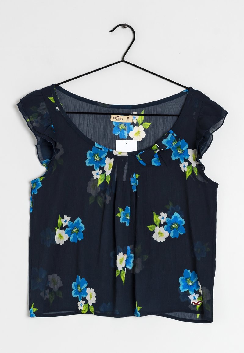 Blouse bleu marine avec manches courtes à volants, décorée de motifs floraux bleus et blancs, suspendue sur un cintre noir.