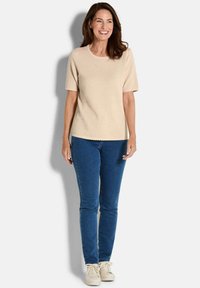Een vrouw die glimlacht, staande, met een beige gestructureerde korte mouwen top, een blauwe jeans en crèmekleurige sneakers tegen een effen witte achtergrond.