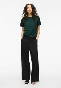 Maglietta di cotone verde scuro a maniche corte abbinata a pantaloni neri larghi. Il modello ha capelli ricci e le mani in tasca.