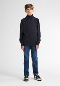 Navyfarbener Rollkragenpullover aus weichem Material, kombiniert mit blauen Jeans und schwarz-weißen Sneakers. Einfaches, tailliertes Design.