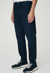 Blugi din denim de culoare albastru închis, cu un croi drept, având un design clasic cu cinci buzunare și detalii de cusătură subtile. Purtați cu adidași negri.