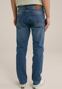 Mėlynos džinsinės kelnės su tiesiu siluetu, turinčios galines kišenes su matoma siuvimo linija, odinę etikėtę ant juosmens ir minimalų dėvėjimą.