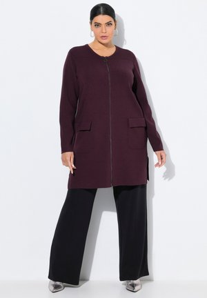 Lange bordeauxrode cardigan met rits, zijsplitten en twee opgestikte zakken, gedragen over zwarte wijde broek en zilveren schoenen.