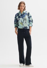 Camicia a fiori blu chiaro con bottoni e accenti blu navy, abbinata a pantaloni alti blu navy scuri. Il modello sta in piedi con le mani nelle tasche e indossa sneakers bianche.